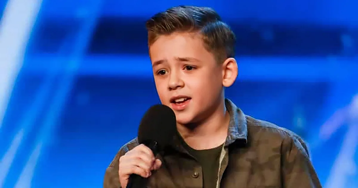 A young boy with short hair holds a microphone, wearing a brown shirt, against a blue stage backdrop.