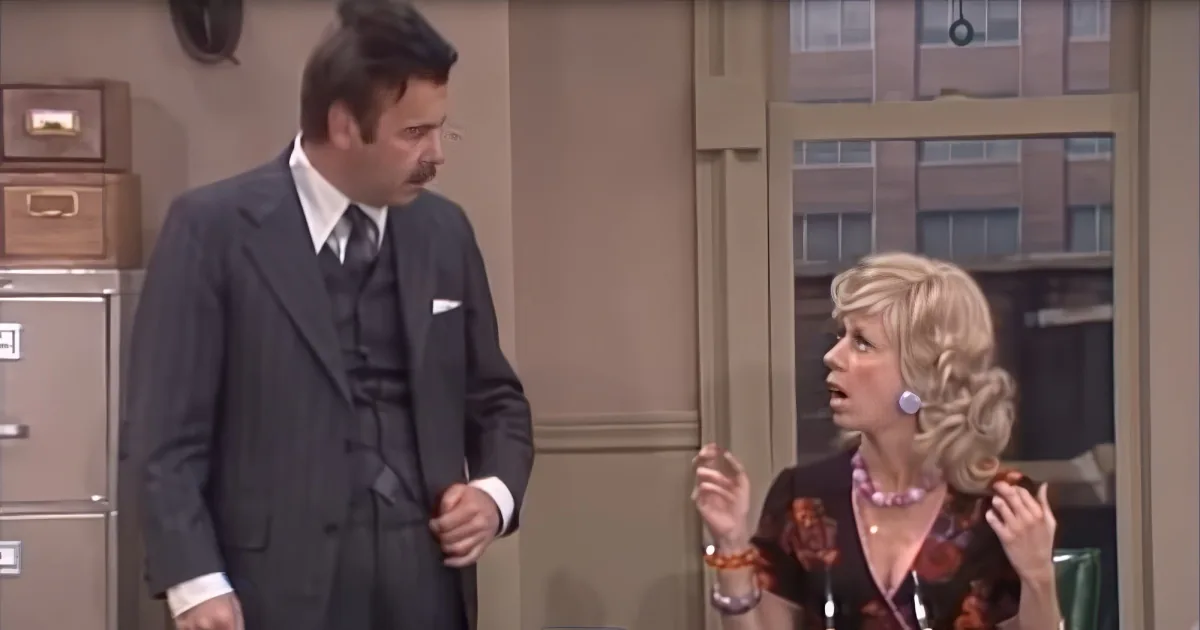 A man in a suit and tie stands in an office, looking down, while a woman in a floral dress reacts with surprise.