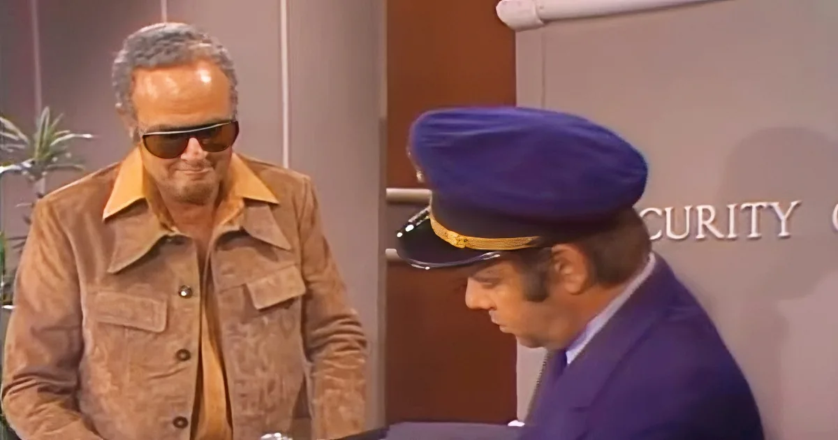 A man in a brown jacket and sunglasses stands beside a security guard in a blue uniform and hat.