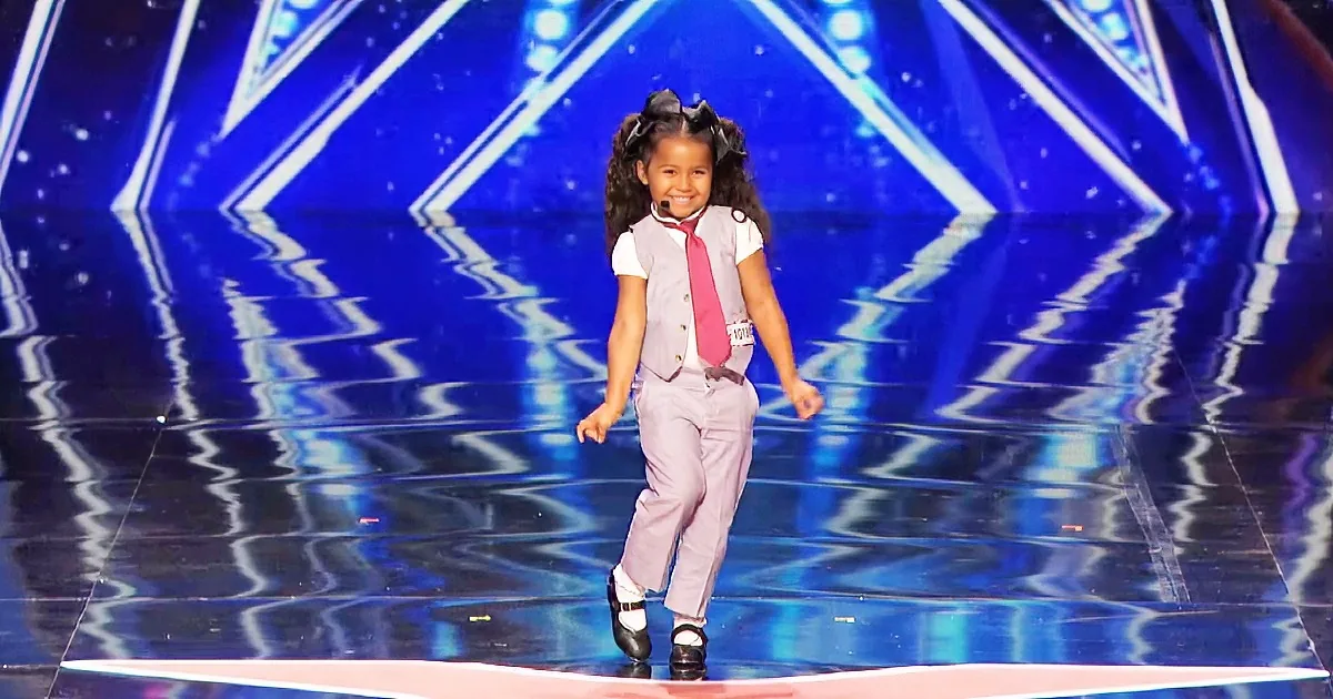 A young girl in a vest and tie walks confidently on a stage with a blue geometric background.
