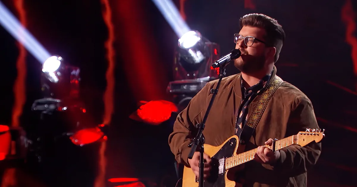 A man in glasses plays a guitar and sings into a microphone on a stage with red lighting.