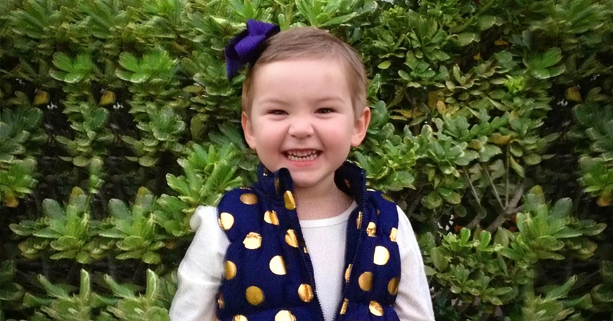 A smiling child with a purple bow in their hair, wearing a navy polka dot vest over a white shirt, standing in front of...