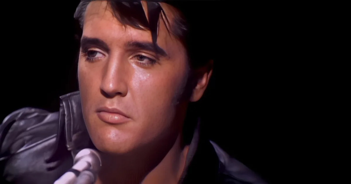A man in a black leather jacket is singing into a microphone, with a dark background.
