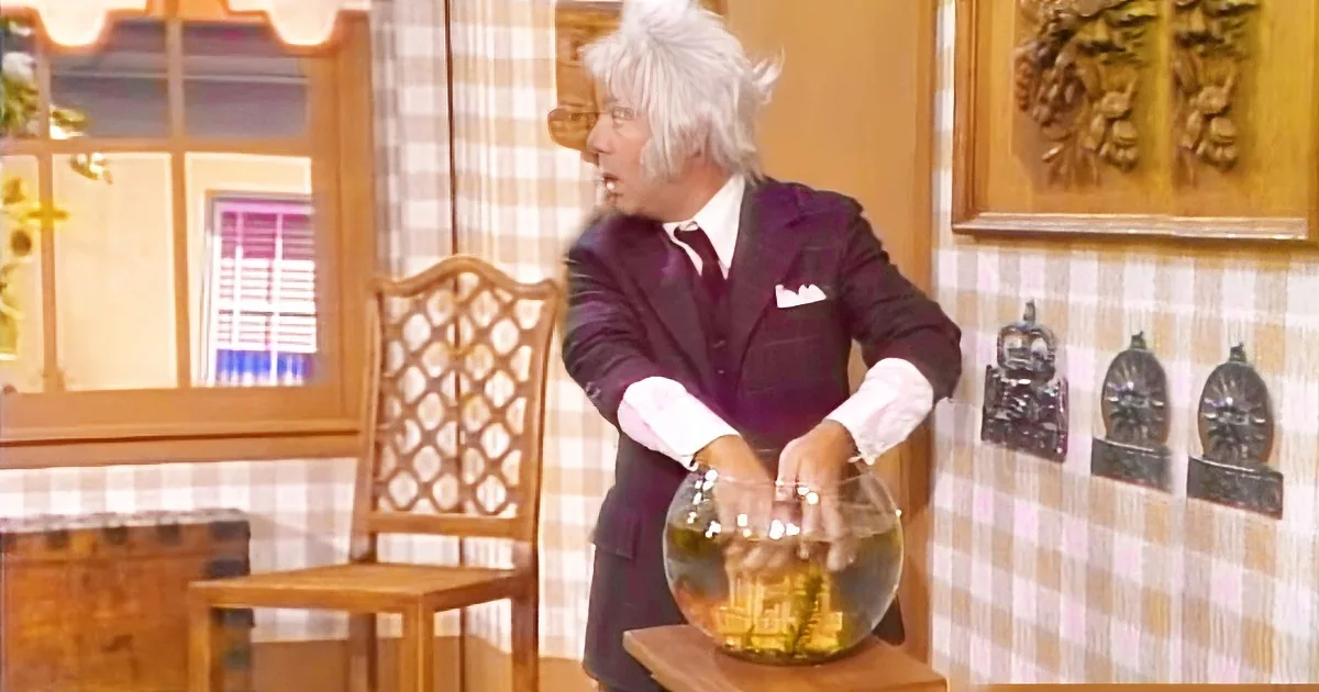 A person in a suit is holding a fishbowl with a fish inside, in a room with checkered wallpaper and wooden furniture.