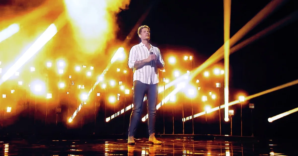 A man stands on a stage with a microphone, surrounded by bright yellow lights and smoke.