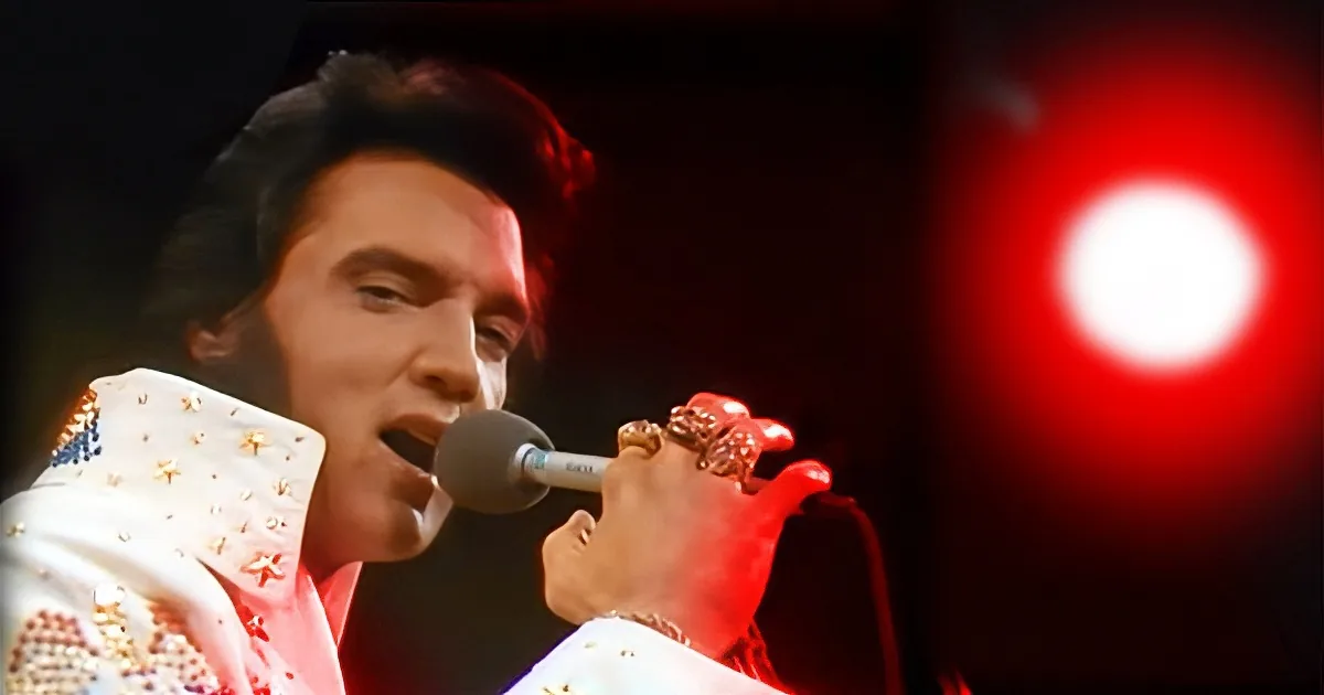 Elvis Presley in a white sequined jacket singing into a microphone with red lighting.