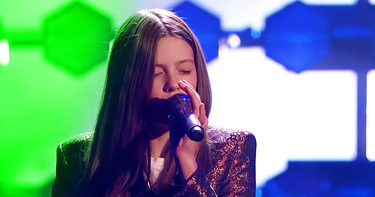 A young girl with long hair sings into a microphone, her eyes closed, in a colorful, illuminated setting.
