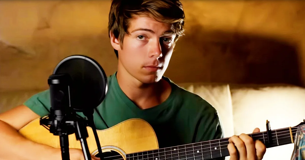 A young man in a green shirt plays an acoustic guitar in front of a microphone.