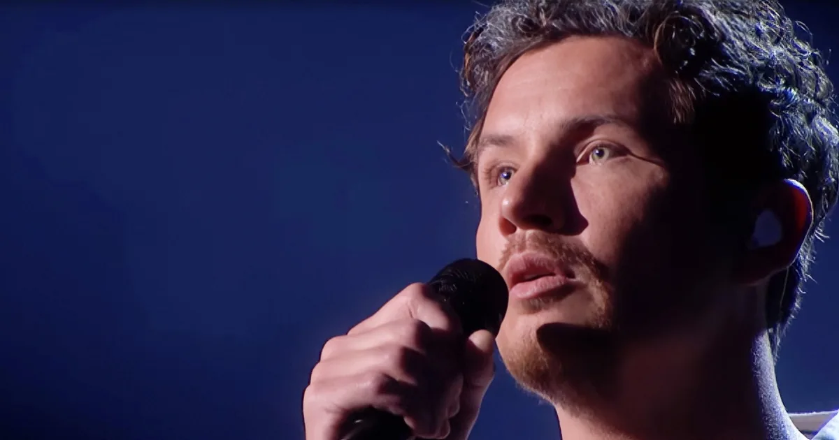 A man with curly hair and a beard holds a microphone, looking upwards, possibly singing or speaking.
