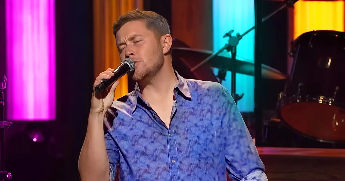 A man in a blue patterned shirt sings into a microphone on stage with colorful lights and a drum set in the background.