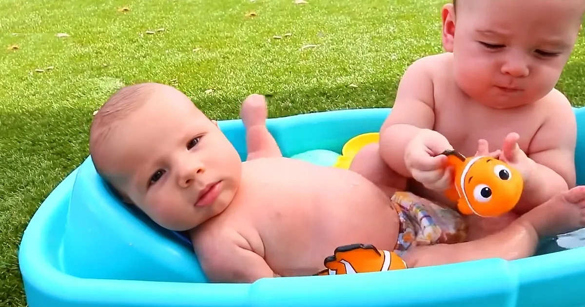 Two babies in a blue plastic tub, one holding a toy fish, on a grassy lawn.