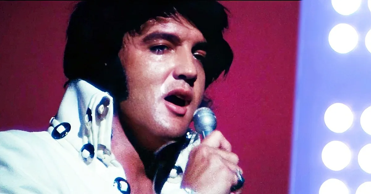 Elvis Presley in a white shirt singing into a microphone with a red background and bright lights.