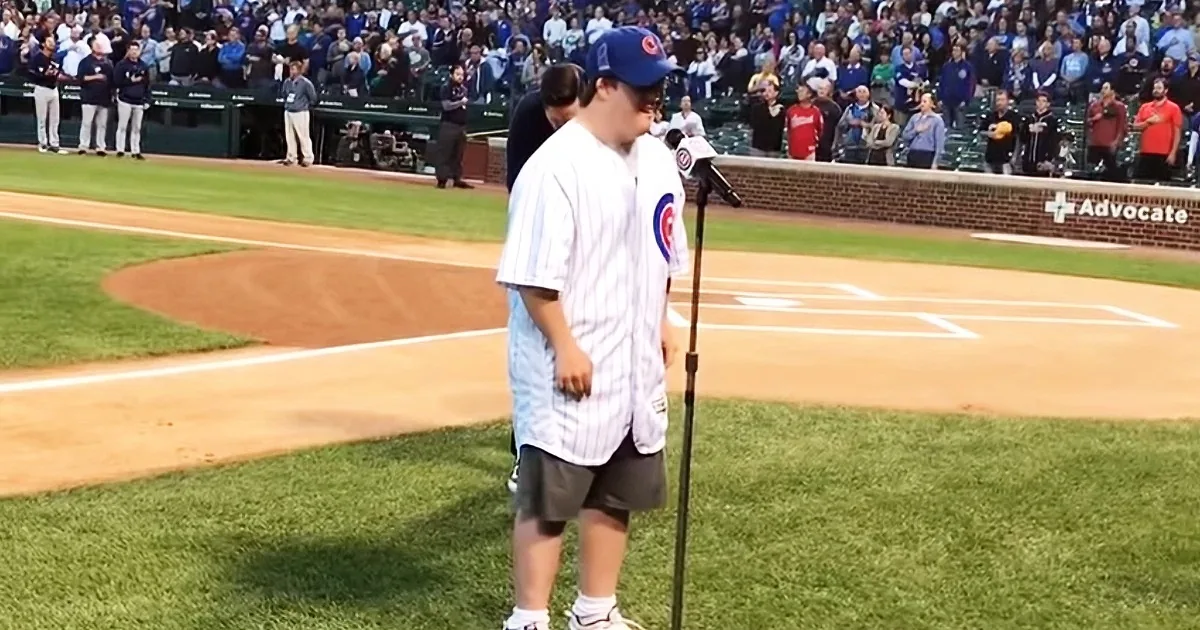 A person in a Cubs uniform stands at a microphone on a baseball field, with a crowd in the background.
