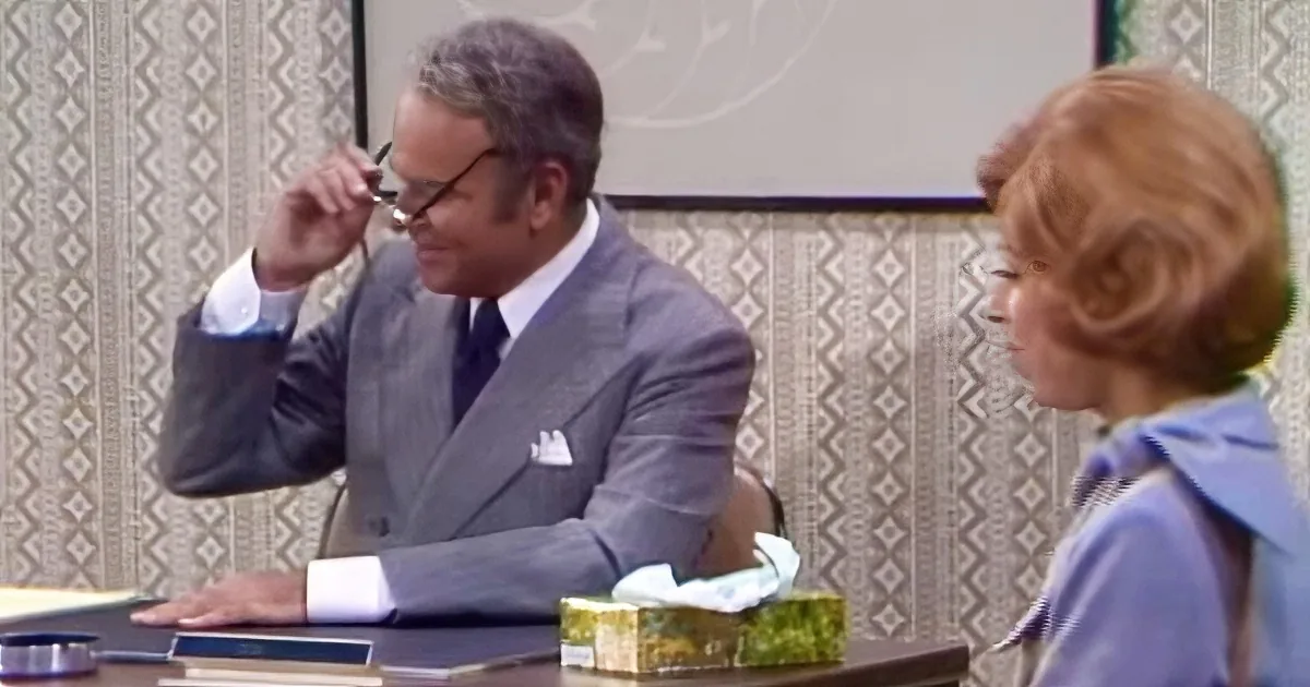 A man in a suit adjusts his glasses while seated at a desk with a tissue box and books.