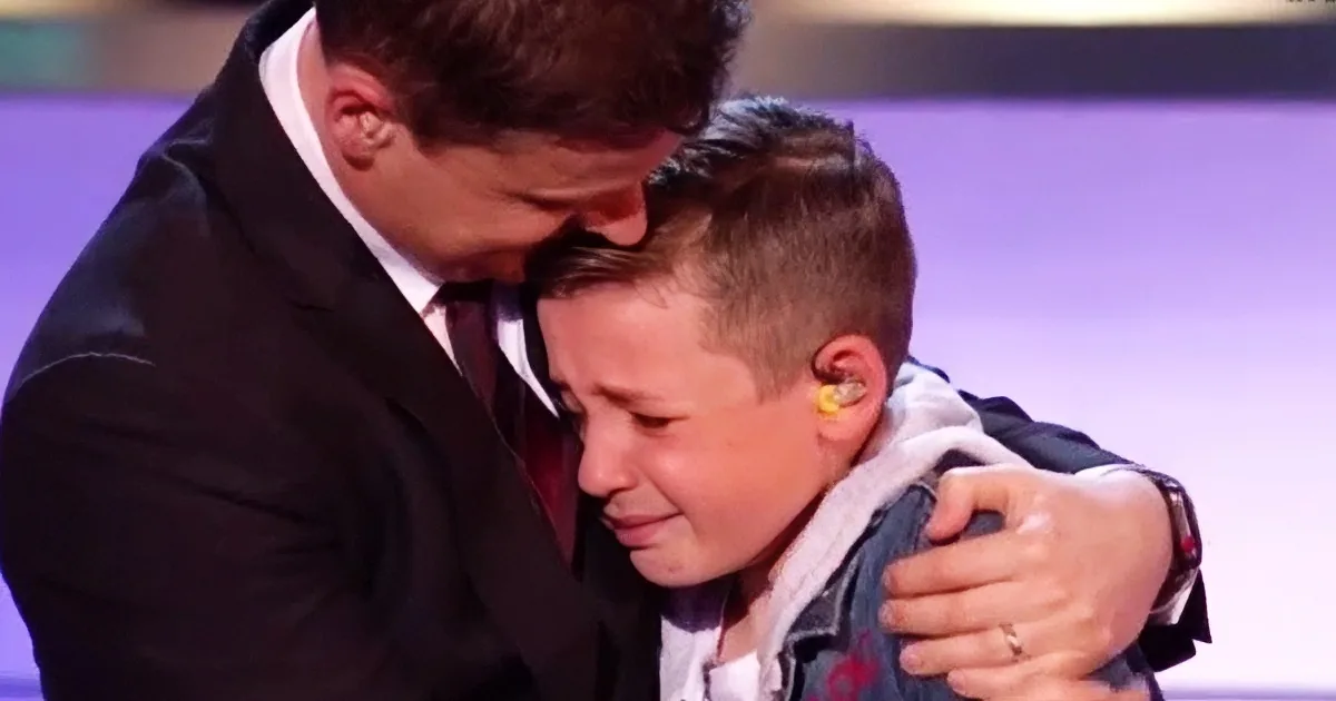 A man in a suit embraces a young boy, both appearing emotional, with the boy wearing a white shirt and a denim jacket.