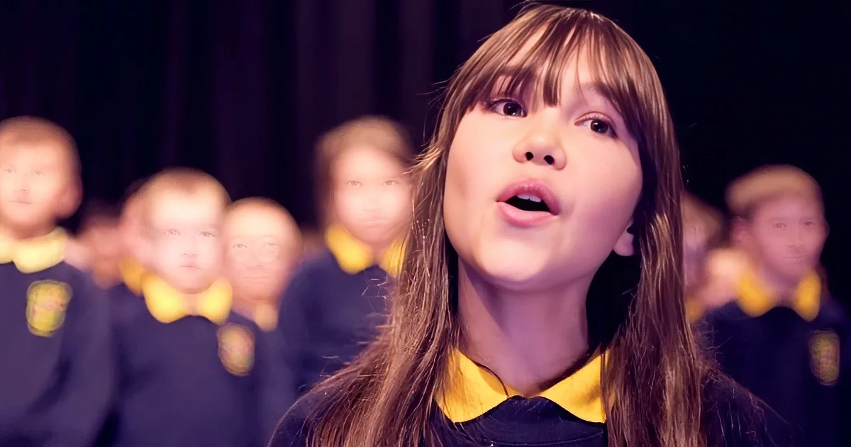 A young girl in a school uniform sings passionately in front of a blurred choir.