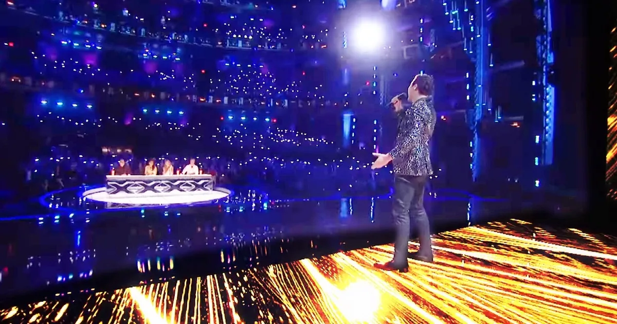 A man in a sequined jacket stands on a stage with bright lights and a reflective floor, singing into a microphone.