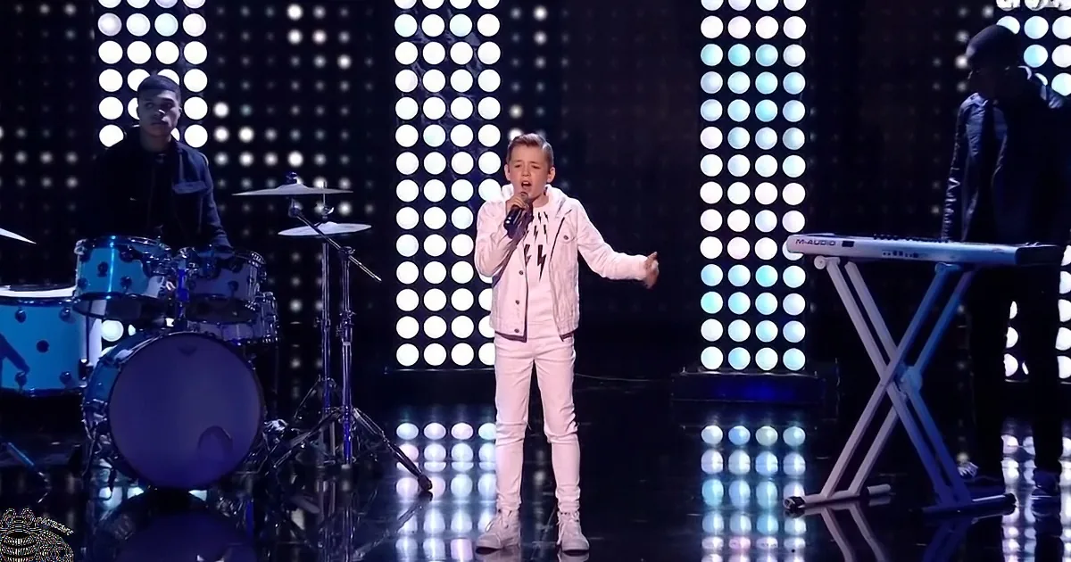 A young boy in white performs on stage with a microphone, surrounded by musicians and bright circular lights.