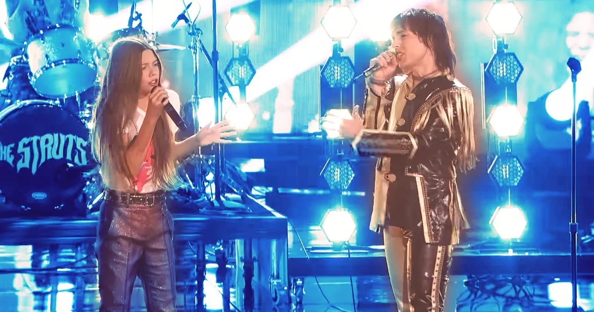 A woman in a white shirt and black pants sings into a microphone on stage with bright lights and a drummer in the background.