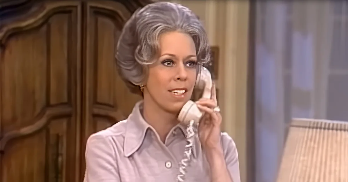 A woman with gray hair in a 1970s-style hairstyle is holding a phone to her ear, wearing a light-colored blouse.