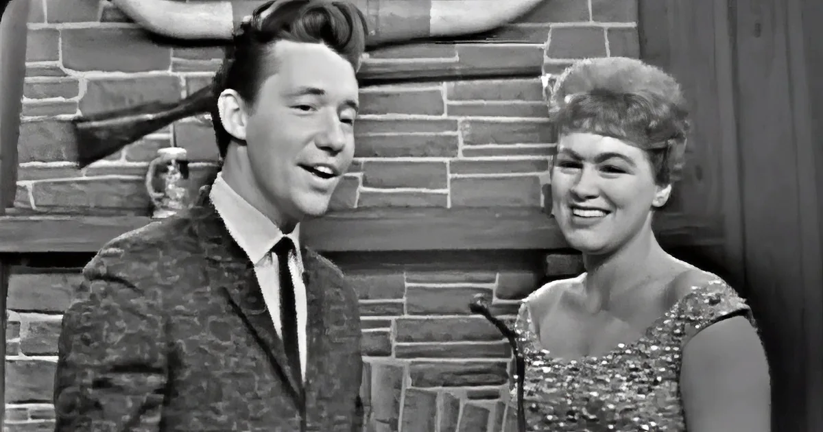 A man in a patterned suit and a woman in a sequined dress smile together in front of a brick wall.