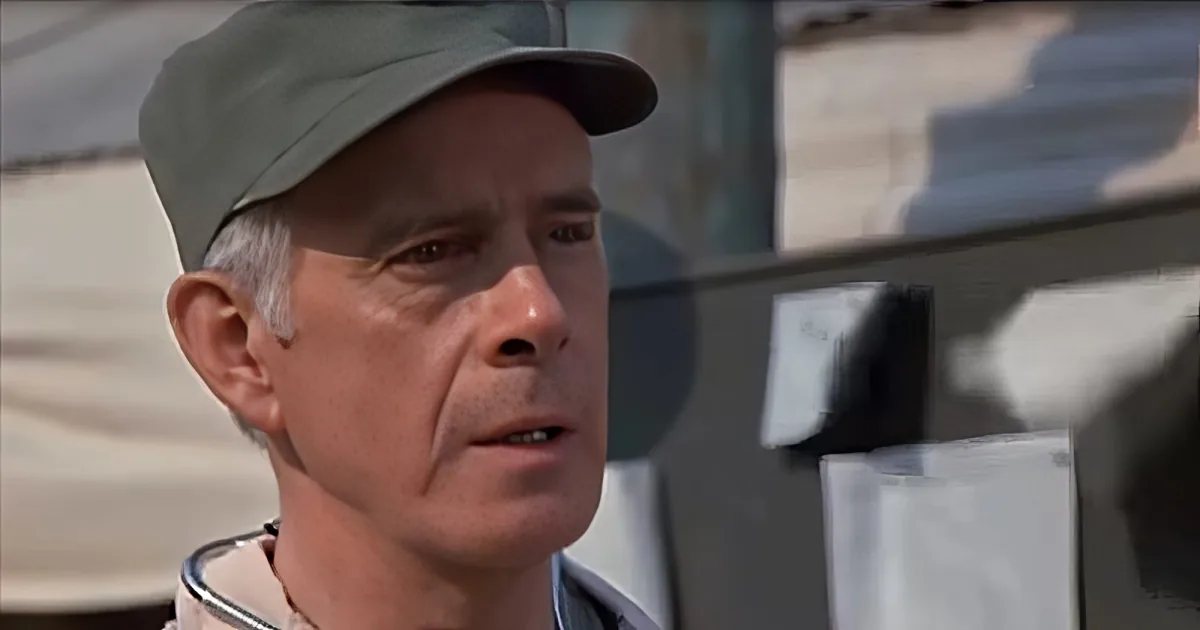 A man wearing a cap and a light-colored shirt is looking to the side with a serious expression.
