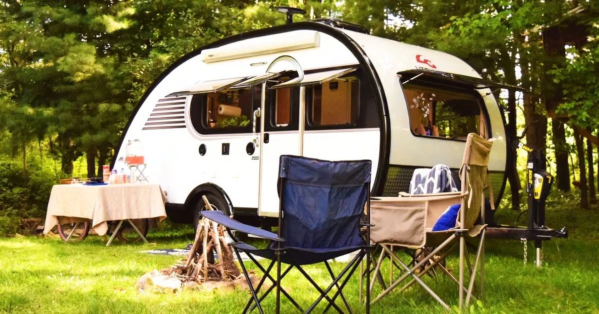 A white teardrop camper with chairs and a table set up in a grassy campsite surrounded by trees.