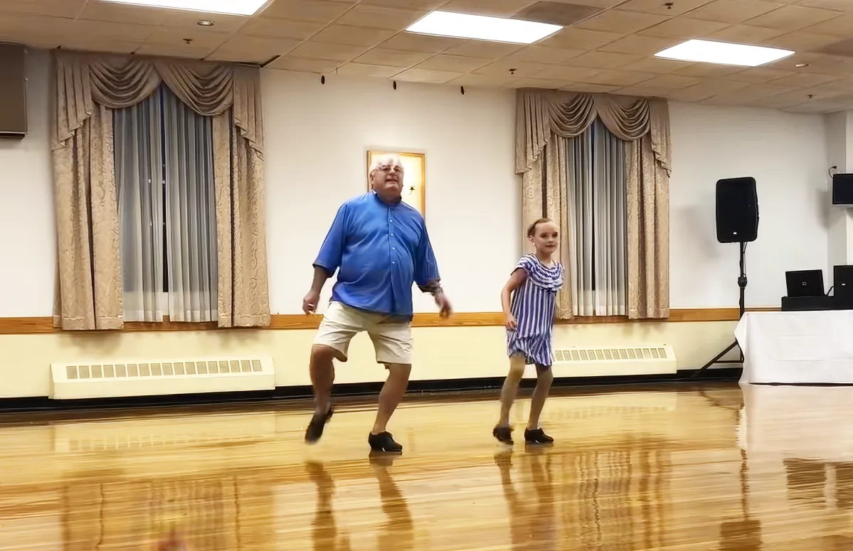 An older man and a young girl dance together on a wooden floor in a room with curtains and a speaker.