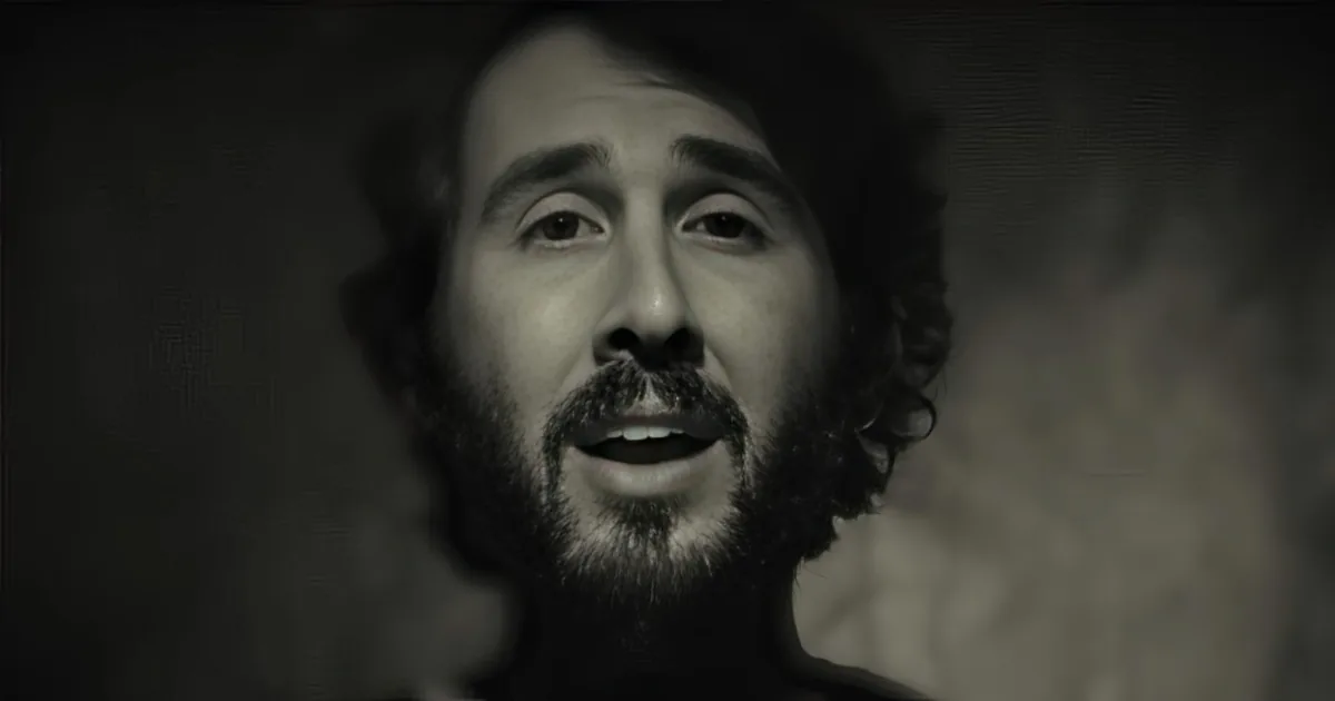 A man with a beard and mustache, wearing a dark shirt, is captured in a close-up shot against a dark background.