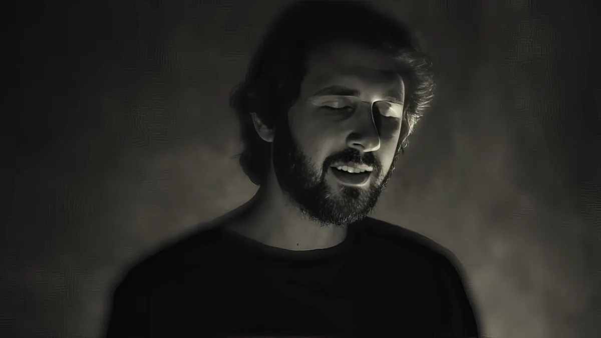 A man with a beard and short hair, wearing a dark shirt, is shown in a dimly lit, monochromatic setting.