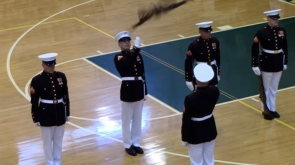A Marine in a black uniform salutes while standing on a basketball court with other Marines.