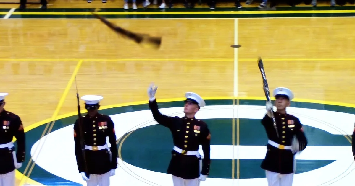 A Marine in uniform raises his hand, holding a baton, with others in formation on a basketball court.