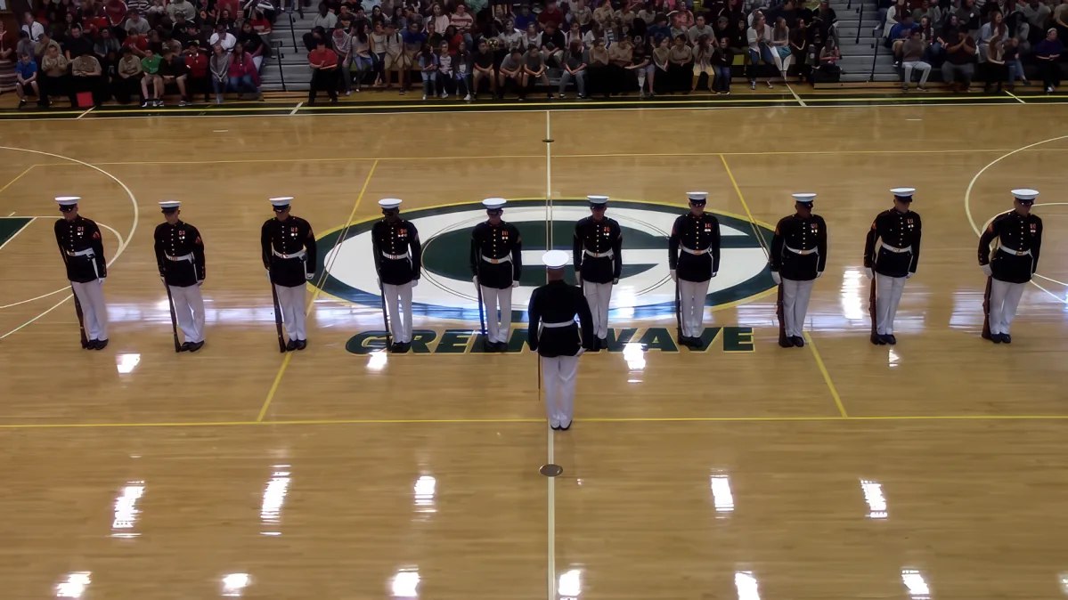 A Marine in uniform stands at attention in front of a line of other Marines on a basketball court.