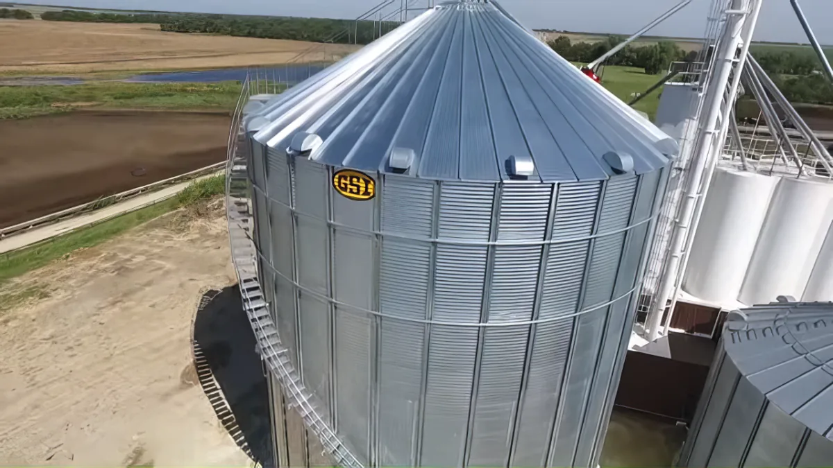 A large metal grain silo with a conical roof stands in a rural landscape, surrounded by fields and trees.