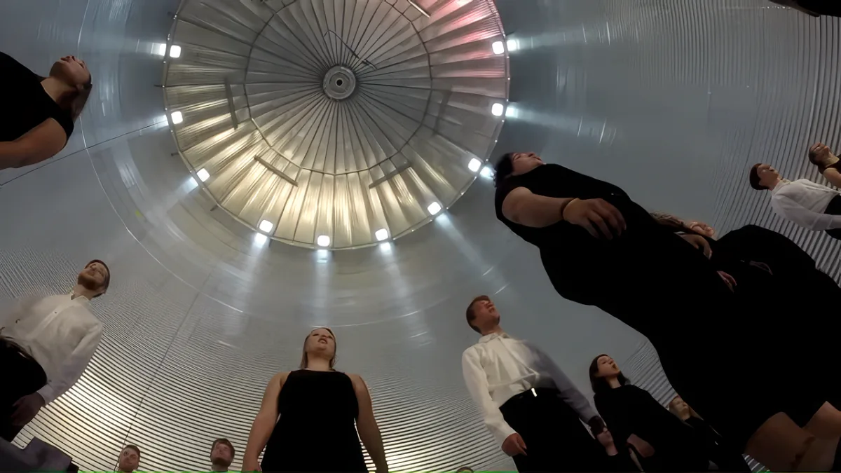 A woman in black stands in a futuristic, metallic room with a large, circular, illuminated structure above her.