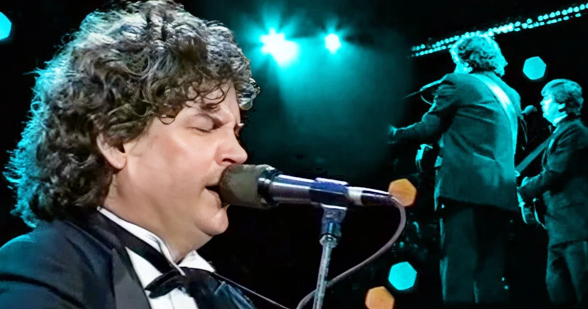 A man with curly hair sings into a microphone on stage, illuminated by blue lights.
