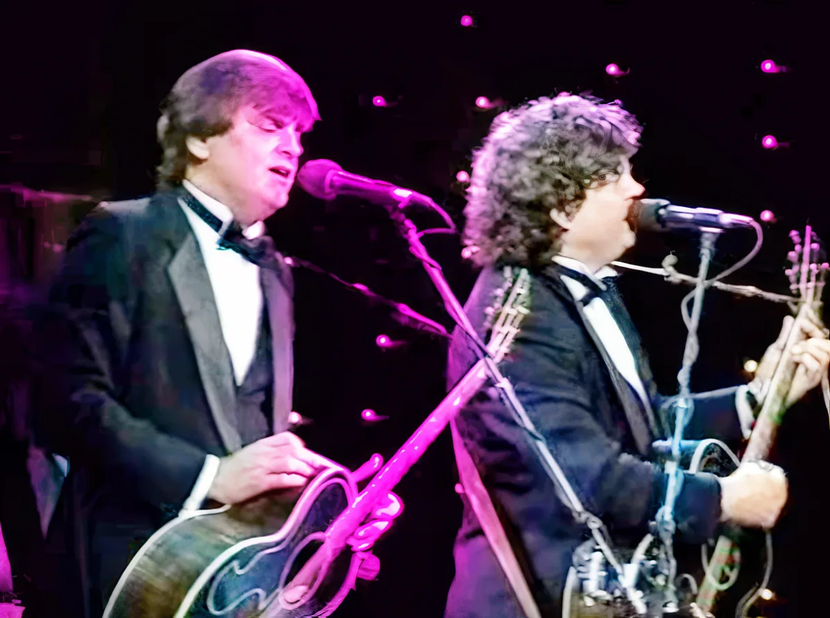 A man in a tuxedo sings into a microphone while another musician plays a stringed instrument.