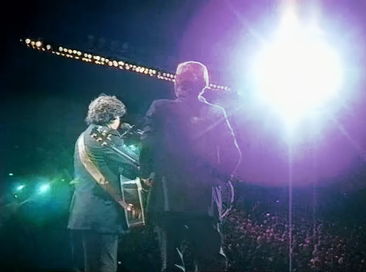 A musician in a dark suit plays guitar under bright stage lights, with another person nearby.