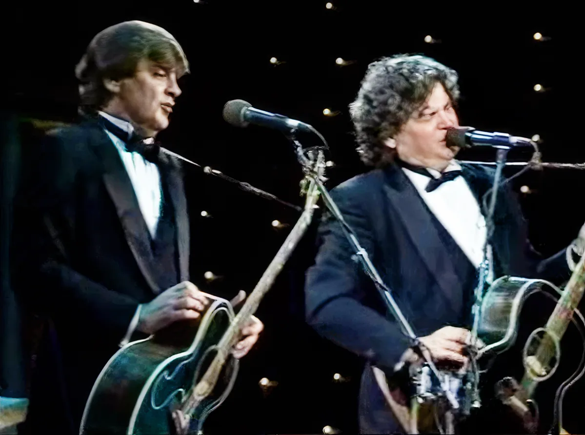 Two men in tuxedos play guitars on stage with microphones and lights behind them.