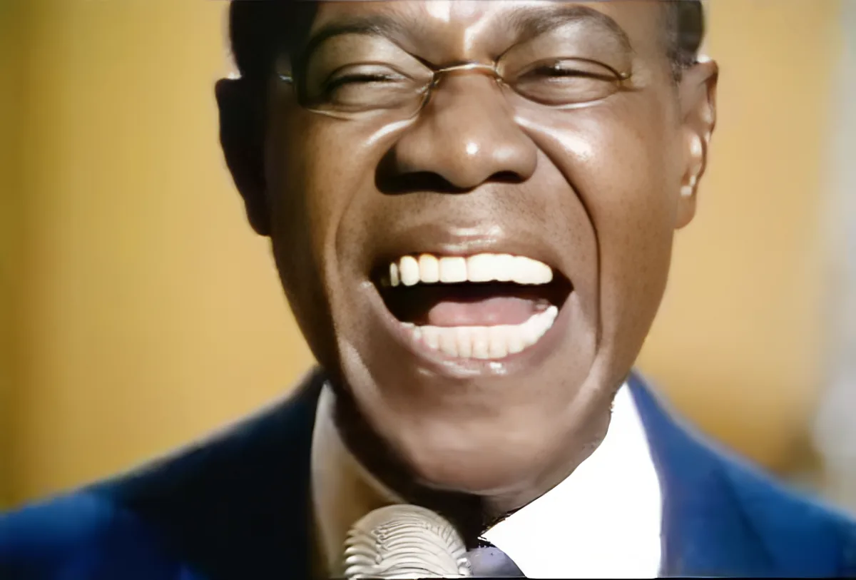 A man in a blue suit sings passionately into a microphone, his mouth wide open.