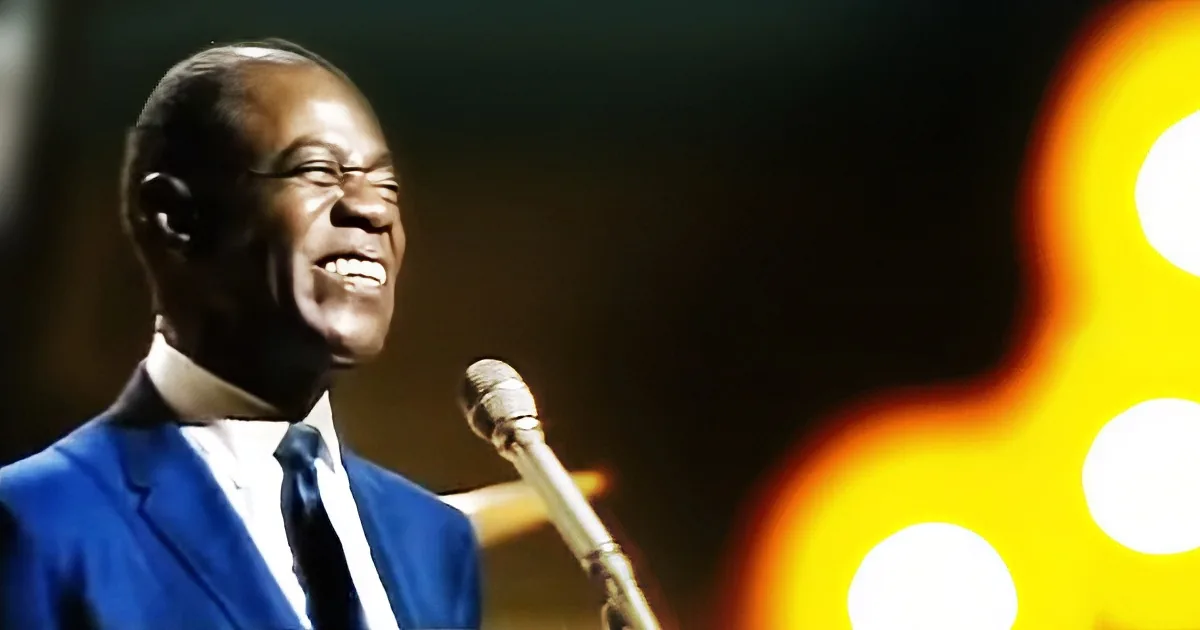 A smiling man in a blue suit sings into a microphone, illuminated by warm stage lights.