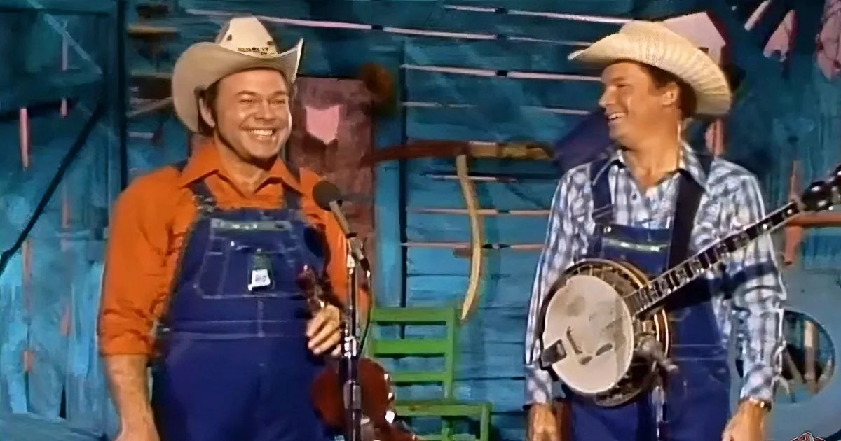Two men in cowboy hats and overalls play instruments on stage, smiling.