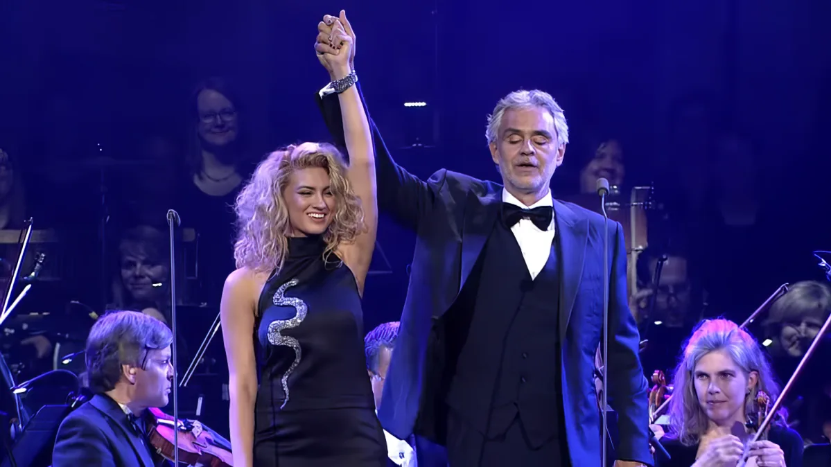 A man in a tuxedo raises his arm in celebration, holding hands with a woman in a black dress, surrounded by an orchestra.