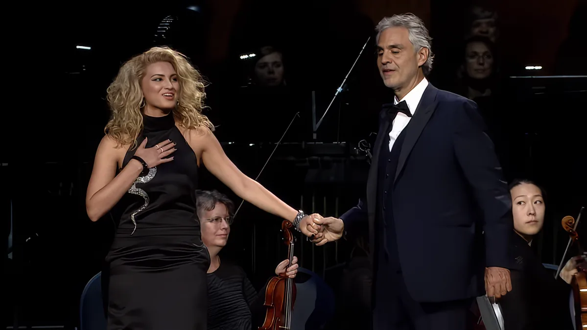 A woman in a black dress shakes hands with a man in a tuxedo on stage, with an orchestra in the background.