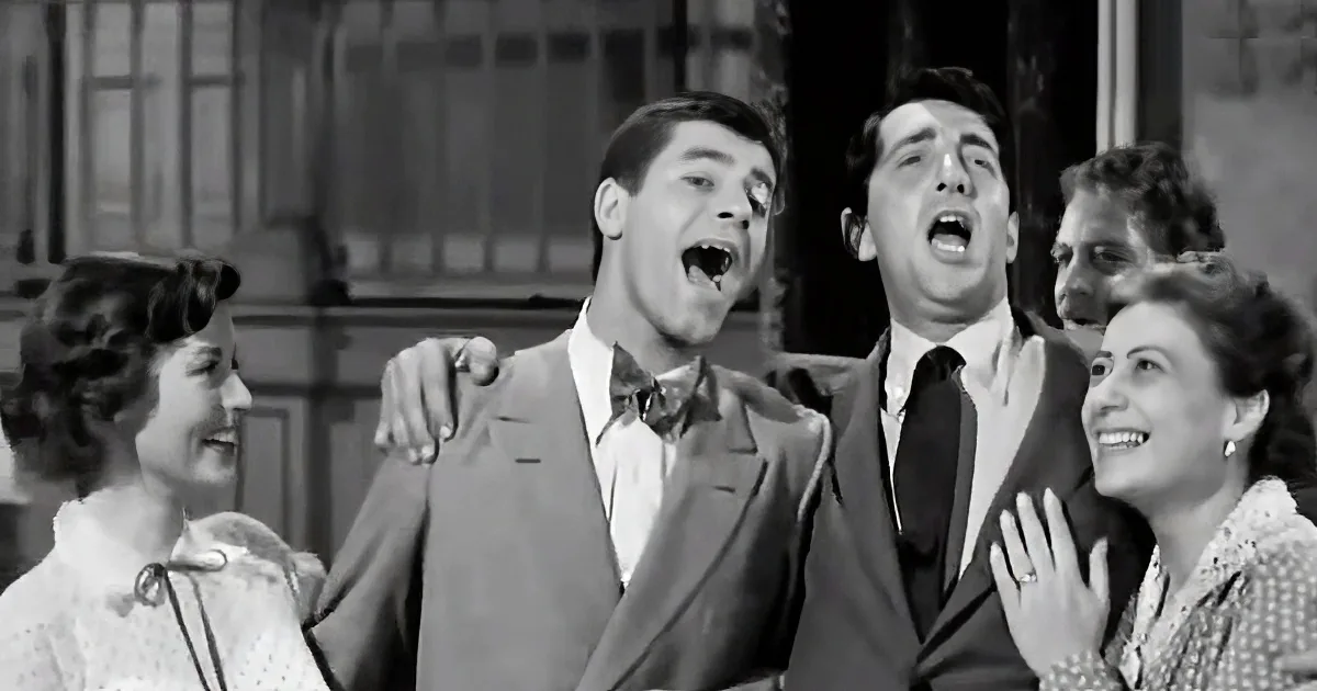 A group of people, including two men in suits, are singing or speaking with their mouths open.