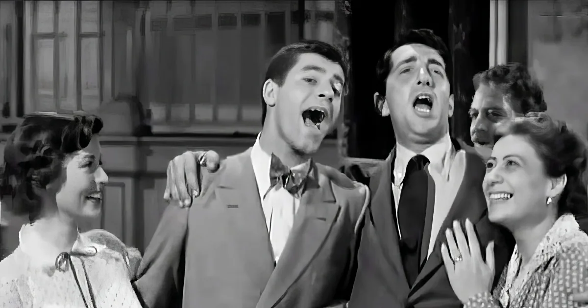 A man in a suit and bow tie is surrounded by others, seemingly in a theatrical or musical performance.