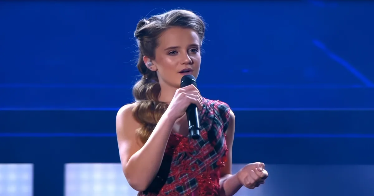 A woman in a red plaid dress holds a microphone, singing on stage with a blue background.
