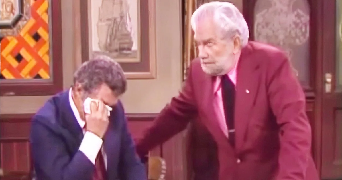 An older man in a pink suit leans over another man in a suit who is holding his face, possibly in distress.