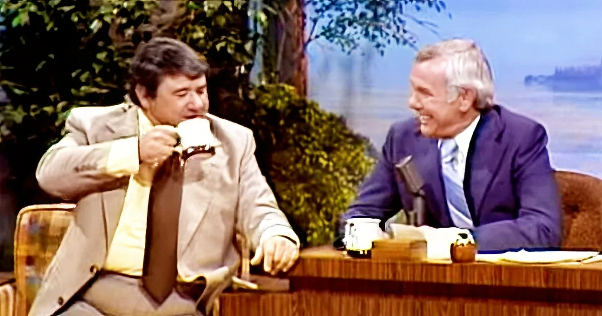 A man in a beige suit drinks from a cup while another man in a blue suit laughs on a talk show set.