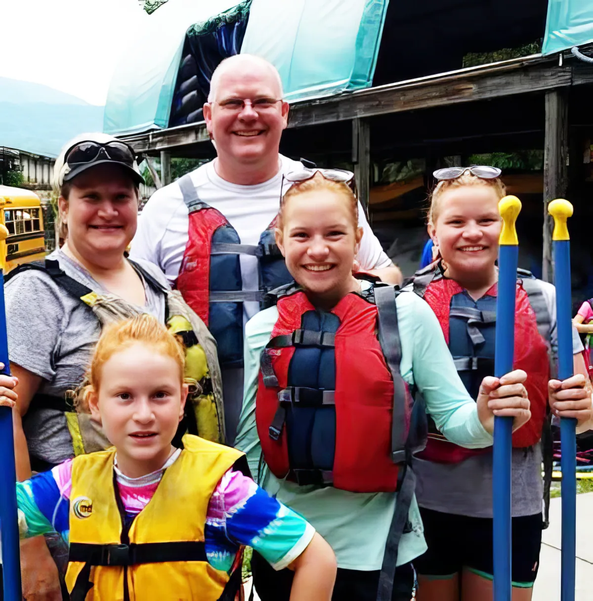 A family wearing life jackets smiles together, holding paddles, in front of a wooden structure.